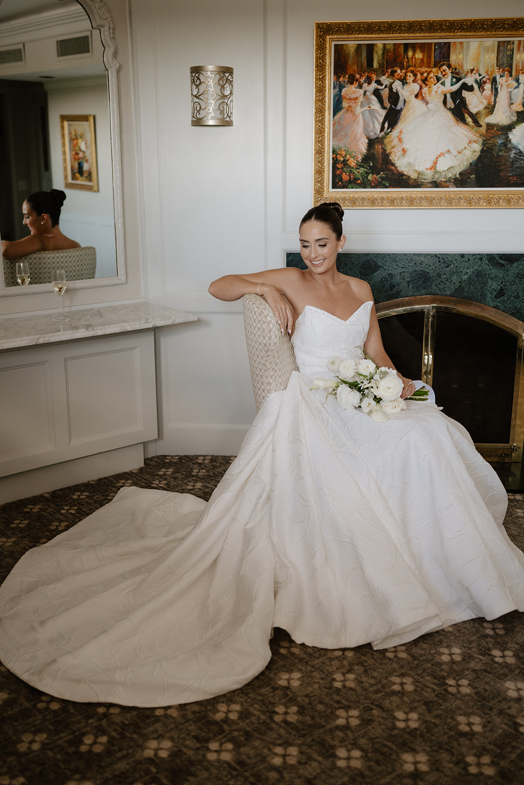 Bride in strapless gown sitting elegantly with her white bouquet in front of a fireplace.